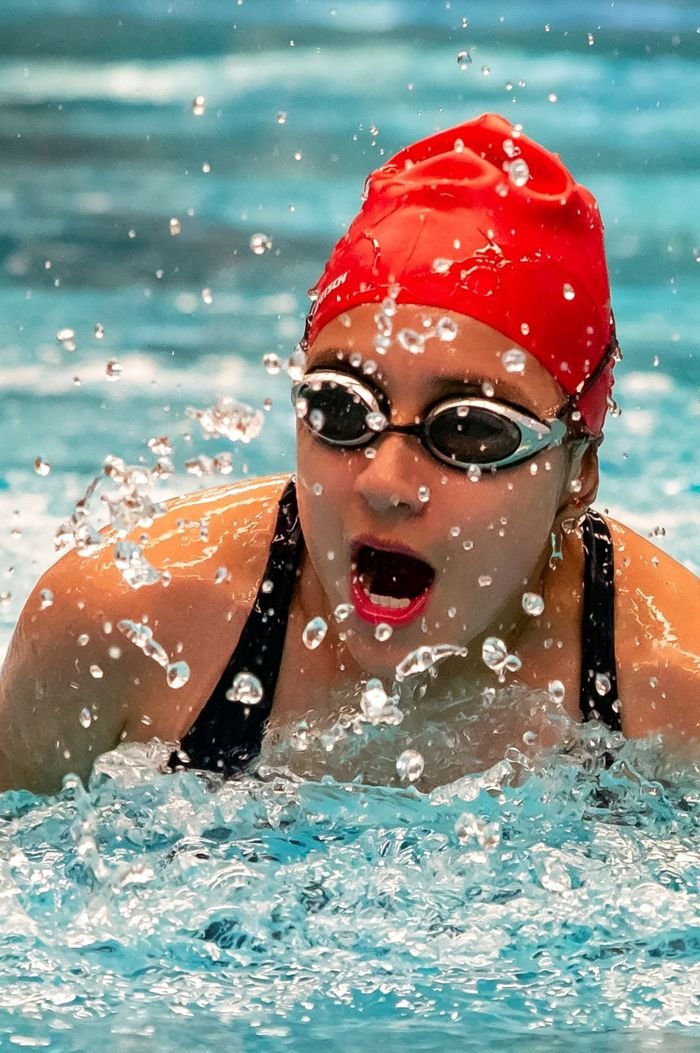 A focused swimmer in a red cap competes in an indoor pool. Water splashes dynamically around.